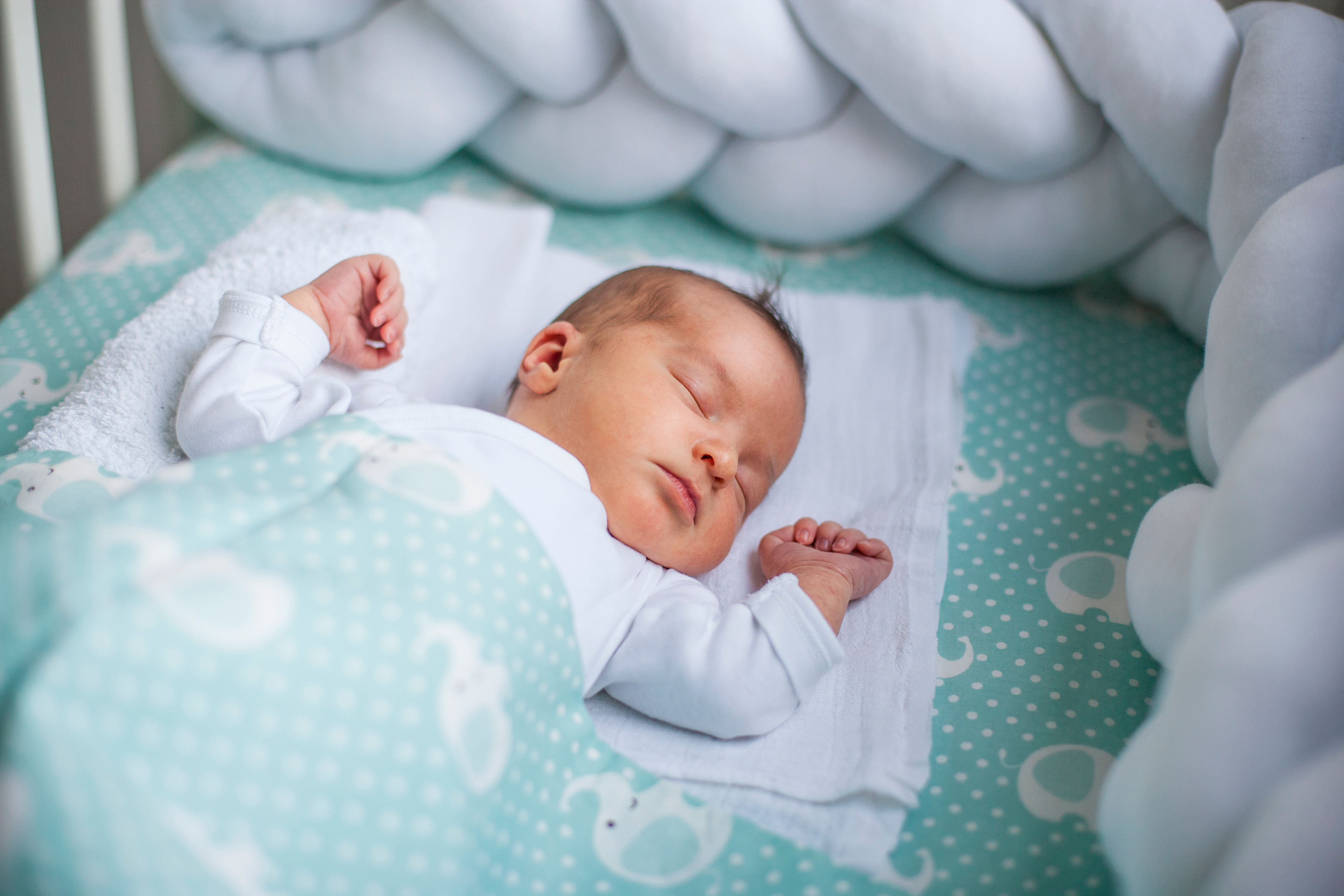 A baby asleep on their back in a crib with braided bumper pads and extra blankets.