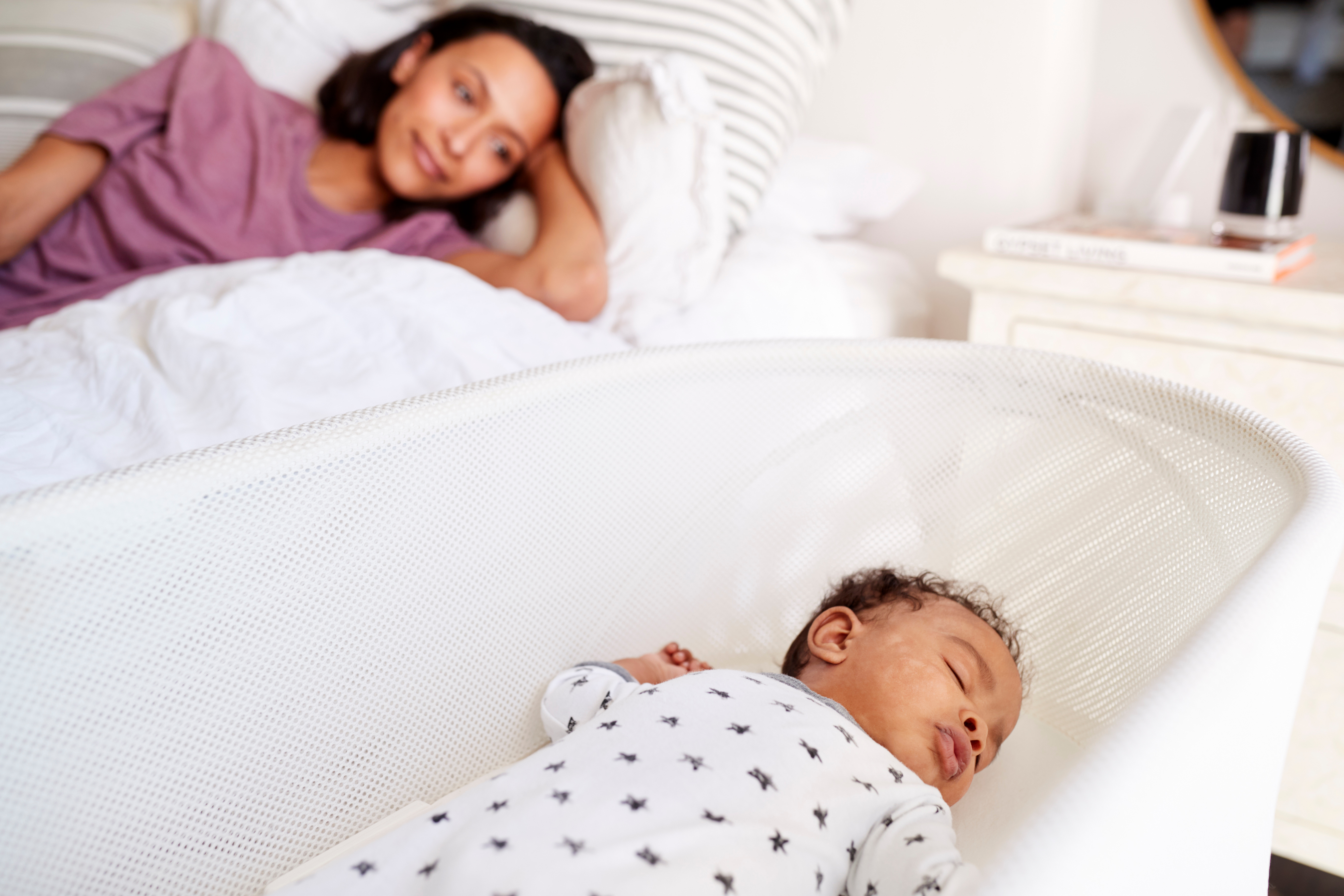 A baby asleep alone on their back in an empty bassinet. A caregiver is nearby.